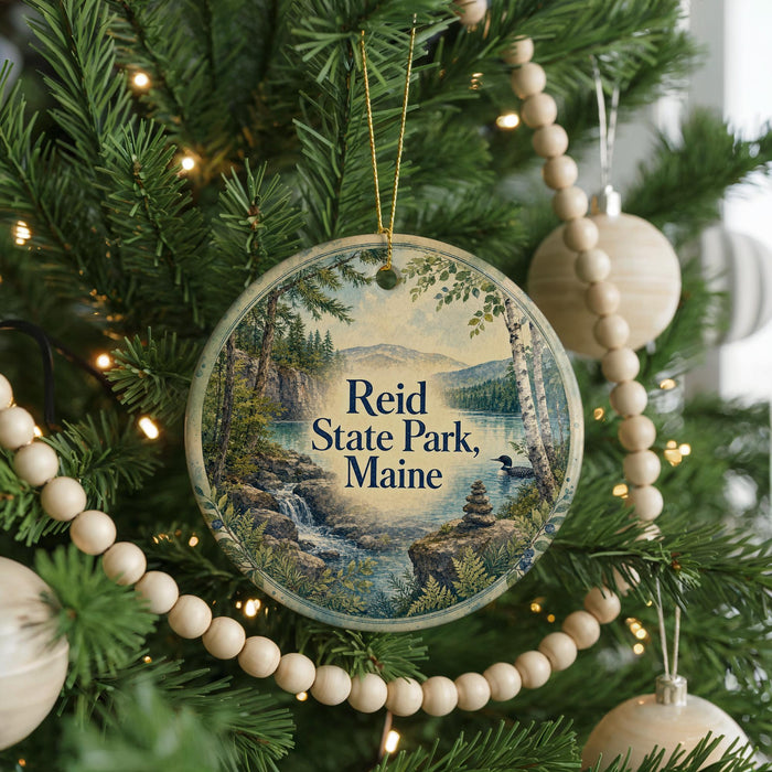 A round ornament on a Christmas tree, featuring a scenic landscape with a waterfall and mountains, with the text "Reid State Park, Maine" displayed on it.