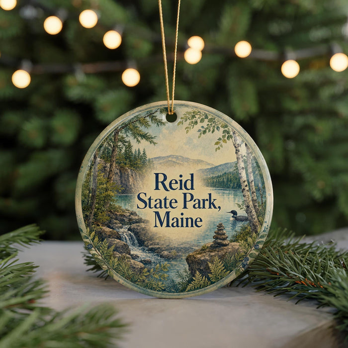 A circular ornament with a scenic landscape of Reid State Park, Maine, featuring a waterfall, pine trees, and mountains.