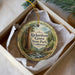 A round ornament with a painting of a forest and a house, hanging from a string inside a box.