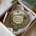 A round ornament with a scenic mountain landscape and a bridge, placed in a box with a white tissue paper lining.