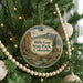 A round ornament hanging on a Christmas tree, featuring a scenic landscape with mountains and a river.