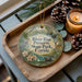A decorative ornament with a nature-themed design, featuring a bird, flowers, and a deer, is displayed on a wooden tray alongside pine cones and a lit candle.