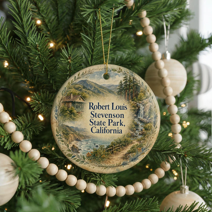 A round ornament on a Christmas tree, featuring a scenic landscape with mountains, a river, and a cabin.