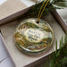 A decorative ornament with a landscape scene featuring a river, trees, and a bridge, placed in a box with pine branches.