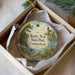 A round ornament with a lighthouse and a scenic landscape, placed in a box with a white tissue paper lining.