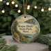 A round ornament with a scenic image of a coastal landscape, featuring a path leading to a cliff overlooking the water, is displayed in front of a blurred background with Christmas lights.