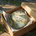 An oval-shaped ornament with a scenic landscape of a coastal area, displayed in a cardboard box with a gold string.
