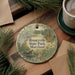 A decorative ornament with a nature scene, featuring a turtle, a deer, and a bird, hangs on a wooden surface alongside a cup of coffee and some pine branches.