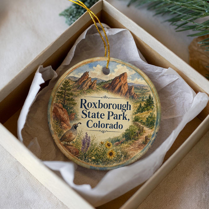 A decorative ornament in a box, featuring a scenic landscape with mountains, a bird, and flowers, with the text "Roxborough State Park, Colorado" printed on it.