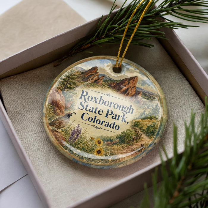 A decorative ornament featuring a scenic landscape of Roxborough State Park, Colorado, with a bird and pine branches.