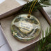 A decorative ornament with a landscape scene featuring a waterfall and pine trees, placed in a box with a pine branch.