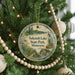 A circular ornament hanging on a Christmas tree, featuring a scenic landscape with a lake, trees, and a bridge.