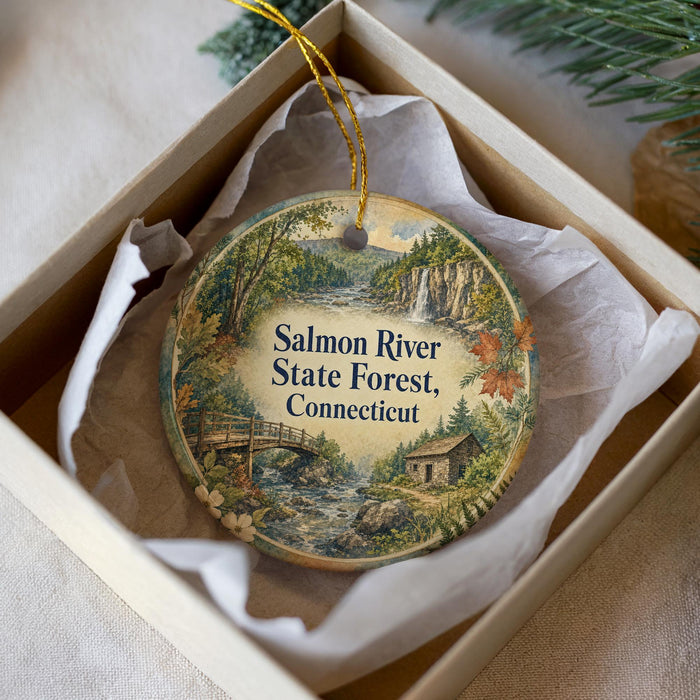 A decorative ornament in a box, featuring a scenic landscape with a river, forest, and bridge.