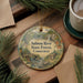 A round ornament with a scenic image of the Salmon River State Forest in Connecticut, surrounded by pine branches and a cup of coffee on a wooden surface.