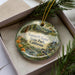 A decorative ornament in a box, featuring a scenic landscape with pine trees, a bird, and a river.