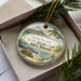 A decorative ornament in a box, featuring a beach scene with palm trees and a mountain in the background.