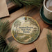A decorative ornament with a bird and palm trees, hanging on a wooden surface, with a cup of coffee and pine branches nearby.