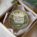 A round ornament with a scenic mountain landscape and a tower, placed inside a box with a white tissue paper lining.