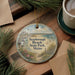 A round ornament with a lighthouse and beach scene, surrounded by pine branches and a cup of coffee on a wooden surface.