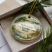 A decorative ornament in a box, featuring a scenic landscape with a lake, trees, and wildlife, including a deer.