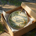 A decorative ornament in a wooden box, featuring a scenic landscape with a lake, trees, and wildlife.