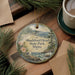 A round ornament with a scenic landscape of Shackford Head State Park, Maine, featuring pine trees, a beach, and a bird, is displayed on a wooden surface alongside a cup of coffee and some papers.