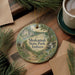 A circular ornament with a scenic landscape of Shokamak State Park, Indiana, hangs on a wooden surface. The ornament is surrounded by pine branches and a cup of coffee, creating a cozy and inviting atmosphere.
