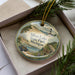 A decorative ornament in a box, featuring a scenic landscape with a lighthouse, trees, and a road.