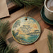 A round ornament with a painting of a bird and a pier, surrounded by pine branches and a cup of coffee on a wooden surface.