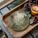 A wooden tray with a decorative ornament featuring a forest scene, pine cones, and a lit candle.