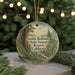A round ornament with a painting of a forest scene, featuring a tall tree and a stream, hangs in front of a Christmas tree with lights.