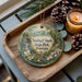 A decorative ornament featuring a scenic image of Smithall Woods State Park, Georgia, with a deer and flowers, placed on a wooden tray alongside pine cones and a lit candle.