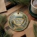 A decorative ornament featuring a scenic landscape of South Mountain State Park, Maryland, with a waterfall and pine trees. The ornament is hanging on a wooden surface, surrounded by pine branches and a cup of coffee.