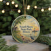 A round ornament with a painting of a lighthouse and a bird, surrounded by greenery and a sunset, hangs on a string.