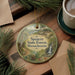 A round ornament with a scenic forest landscape and a waterfall, featuring the text "Spenser State Forest, Massachusetts" on it, is displayed on a wooden surface alongside a cup of coffee and some pine branches.