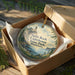 A round ornament in a box, featuring a lighthouse and a scenic landscape, with the text "Split Rock Lighthouse State Park, Minnesota" printed on it.