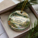 A decorative ornament in a box, featuring a scenic landscape with a pond, pine trees, and mountains.