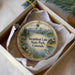 A decorative ornament in a box, featuring a scenic landscape with mountains, a lake, and a cabin, along with a moose and a fish.