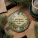 A decorative ornament with a scenic landscape of Stephens State Forest, Iowa, featuring a castle, trees, and a river. The ornament is hanging on a wooden surface, surrounded by pine branches and a cup of coffee.