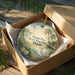 A circular ornament in a wooden box, featuring a beach scene with palm trees, a bird, and seashells.