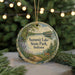 A decorative ornament featuring a scenic landscape with a lake, trees, and a deer, hanging in front of a Christmas tree with lights.