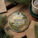 A decorative ornament with a scenic landscape of Susquehanna State Park, Maryland, featuring a river, trees, and mountains. The ornament is hanging from a string and is placed on a wooden surface, surrounded by pine branches and a cup of coffee.
