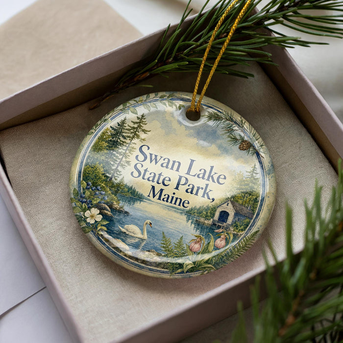 A decorative ornament in a box, featuring a scenic image of Swan Lake State Park in Maine.