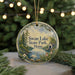A round ornament with a painting of a lake and trees, featuring a swan and a house, hangs in front of a Christmas tree with lights.