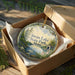 A decorative ornament in a wooden box, featuring a scenic landscape with a lake, trees, and a swan.