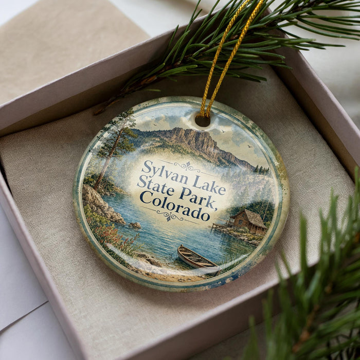 A decorative ornament in a box, featuring a scenic landscape with mountains, water, and a small boat.