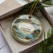 A decorative ornament in a box, featuring a scenic landscape with mountains, water, and a small boat.