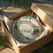 A round ornament in a box, featuring a scenic landscape with mountains, water, and a boat, is displayed on a wooden surface.