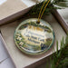 A decorative ornament featuring a scenic image of Tahquamenon Falls State Park, Michigan, with a black bear in the foreground.