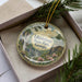 A decorative ornament in a box, featuring a scenic landscape with a waterfall and pine trees.
